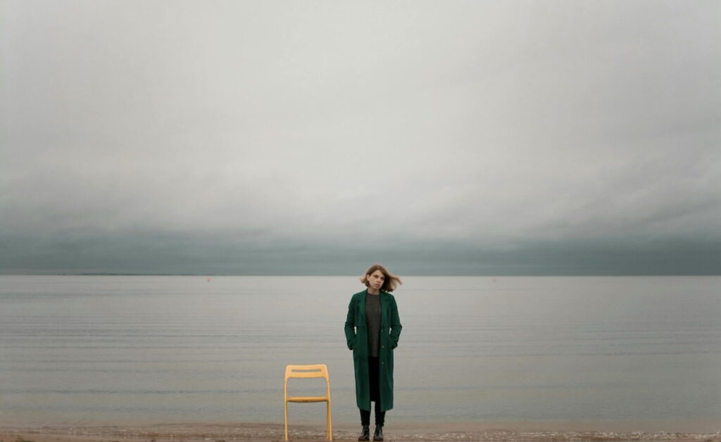 Eine Frau neben einem Stuhl am Meer mit grauem Himmel.